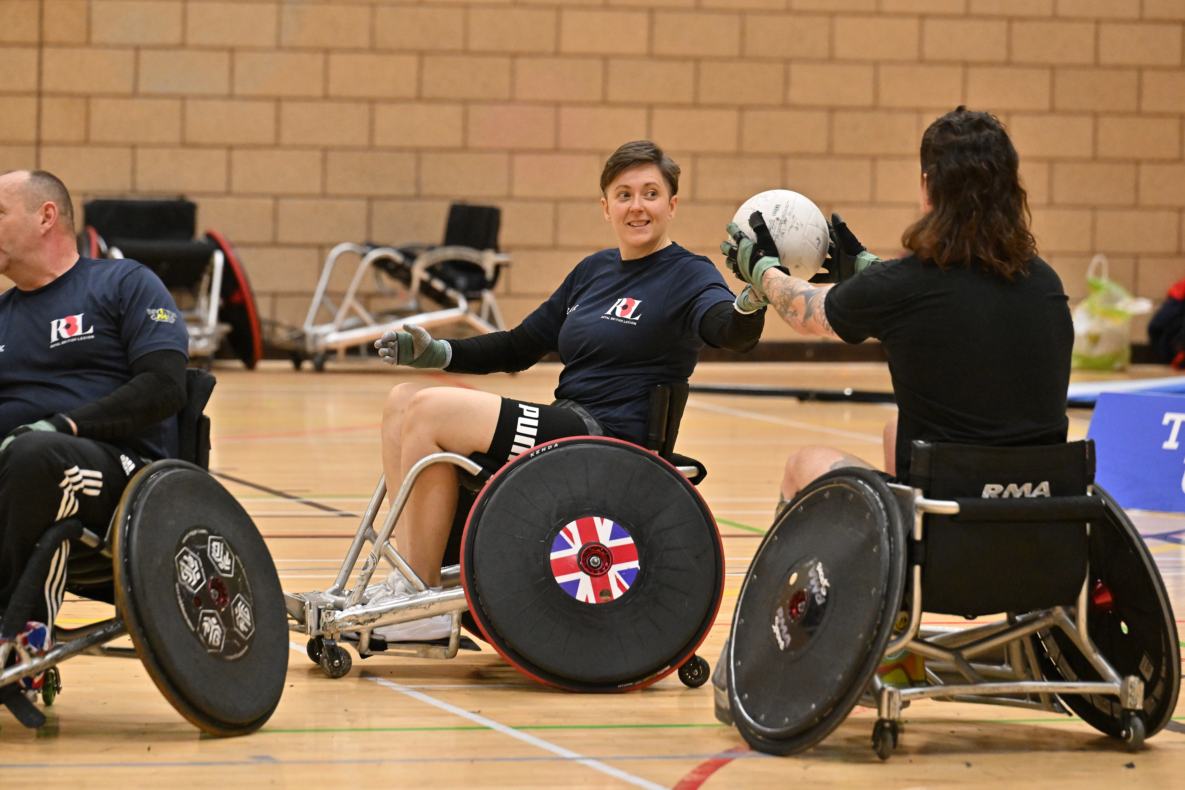 Liz on a basketball court playing wheelchair basketball. She is in a wheelchair, about to take the ball another wheelchair user is passing to her..