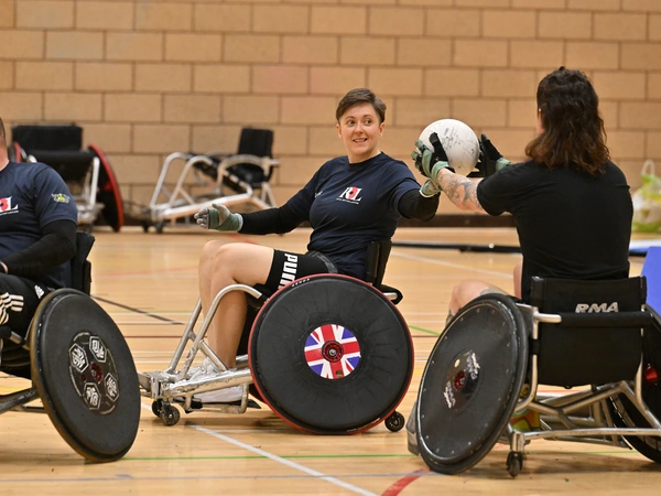 Liz on a basketball court playing wheelchair basketball. She is in a wheelchair, about to take the ball another wheelchair user is passing to her..