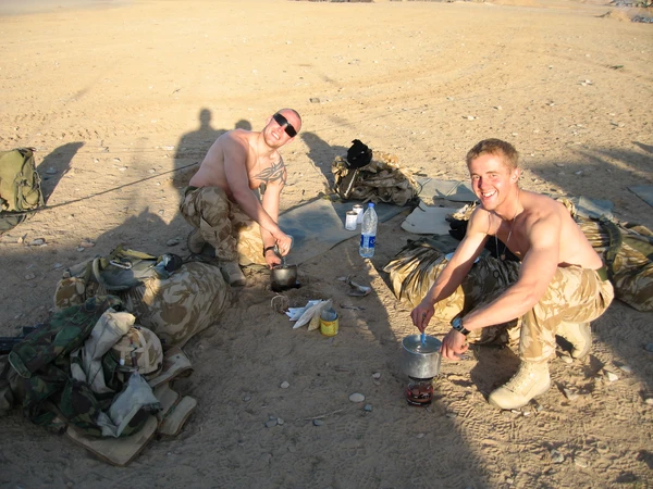Charlie on tour. He is crouched over some cooking utensils together with another colleague. They are wearing combat gear and are in the middle of a dusty road.