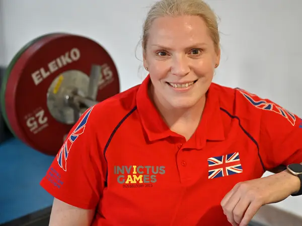 Close up photo of Amanda in a red Invictus Games Team UK polo shirt. We can see some weights on a barbell behind her. She is leaning against the barbell. She is smiling at the camera.