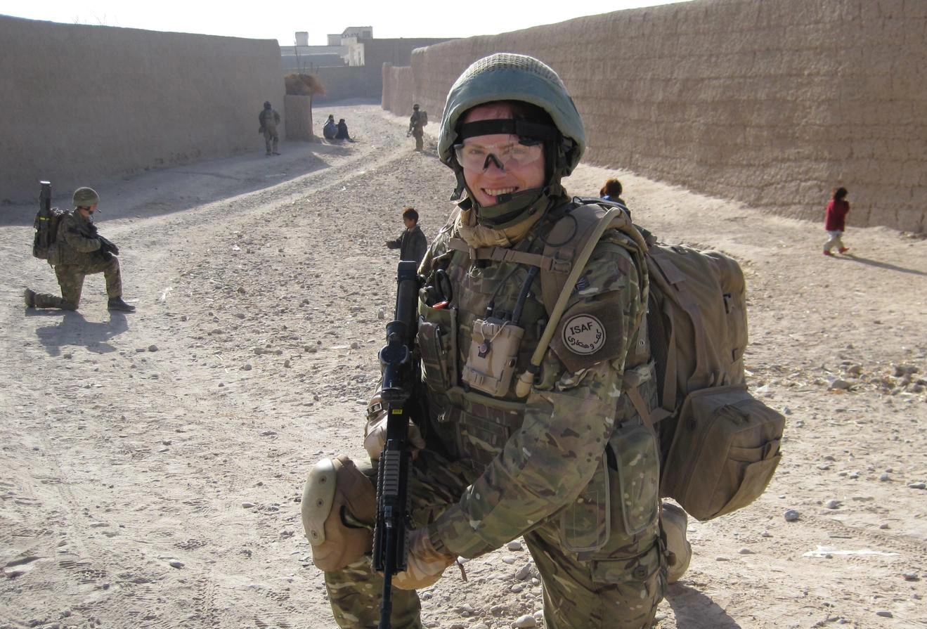 Amanda in full combat gear holding a gun, she is kneeling i the middle of a dirt road. Behind her we can see more soldiers.