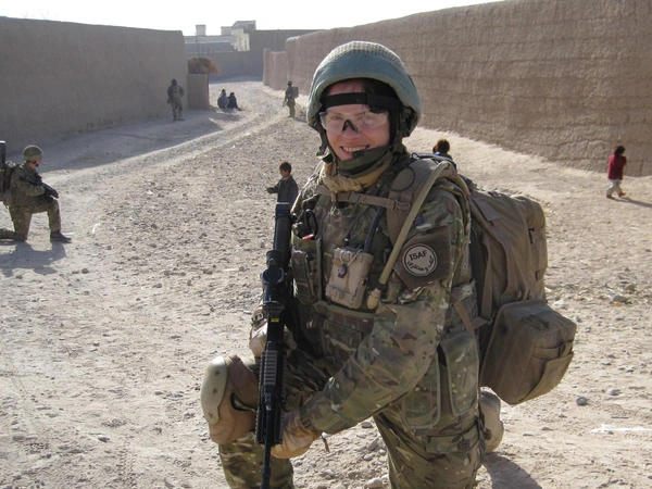 Amanda in full combat gear holding a gun, she is kneeling i the middle of a dirt road. Behind her we can see more soldiers.