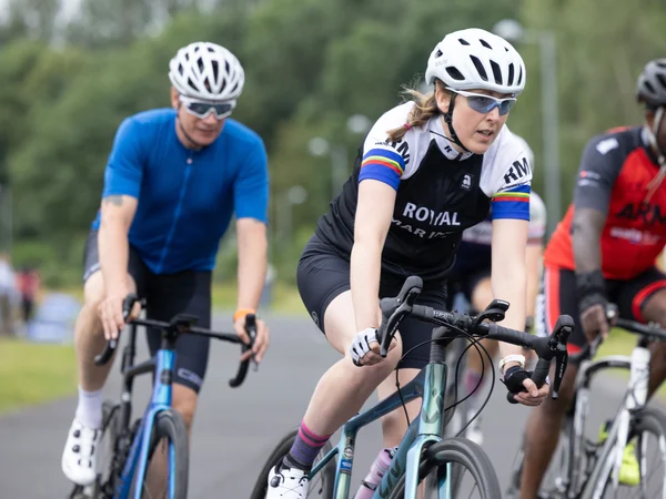 Becky cycling alongside other cyclists on a track at an Invictus training camp. She is wearing full cycling gear and has a concentrated expression on her face.
