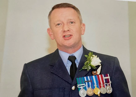 A photo of Al standing in front of a white wall. He is wearing his RAF uniform with medals pinned to it. He also has a white rose pinned to his lapel. He looks to be in mid speech.