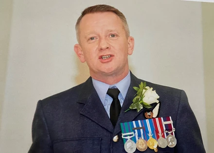 Dyer3 A photo of Al standing in front of a white wall. He is wearing his RAF uniform with medals pinned to it. He also has a white rose pinned to his lapel. He looks to be in mid speech.