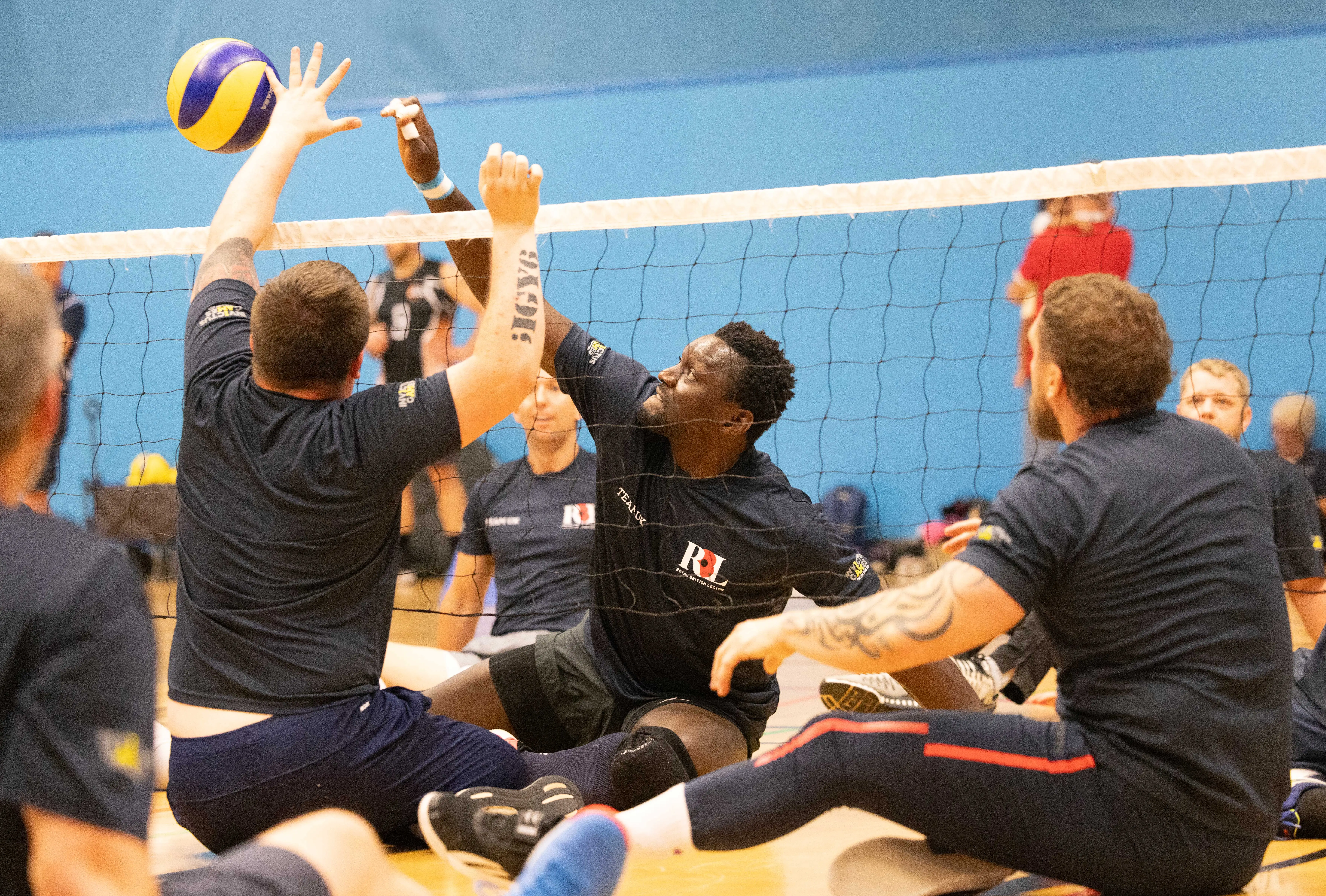Emmanuel during a game of sitting volleyball. He is photographed in action just as he's about to hit the ball over the net. He has a fierce expression on his face. Around him we can see other sitting volleyball players. Everyone is wearing Team UK kit.