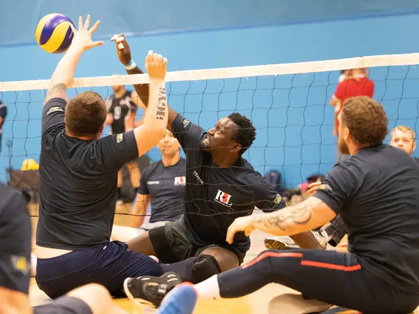 Emmanuel during a game of sitting volleyball. He is photographed in action just as he's about to hit the ball over the net. He has a fierce expression on his face. Around him we can see other sitting volleyball players. Everyone is wearing Team UK kit.