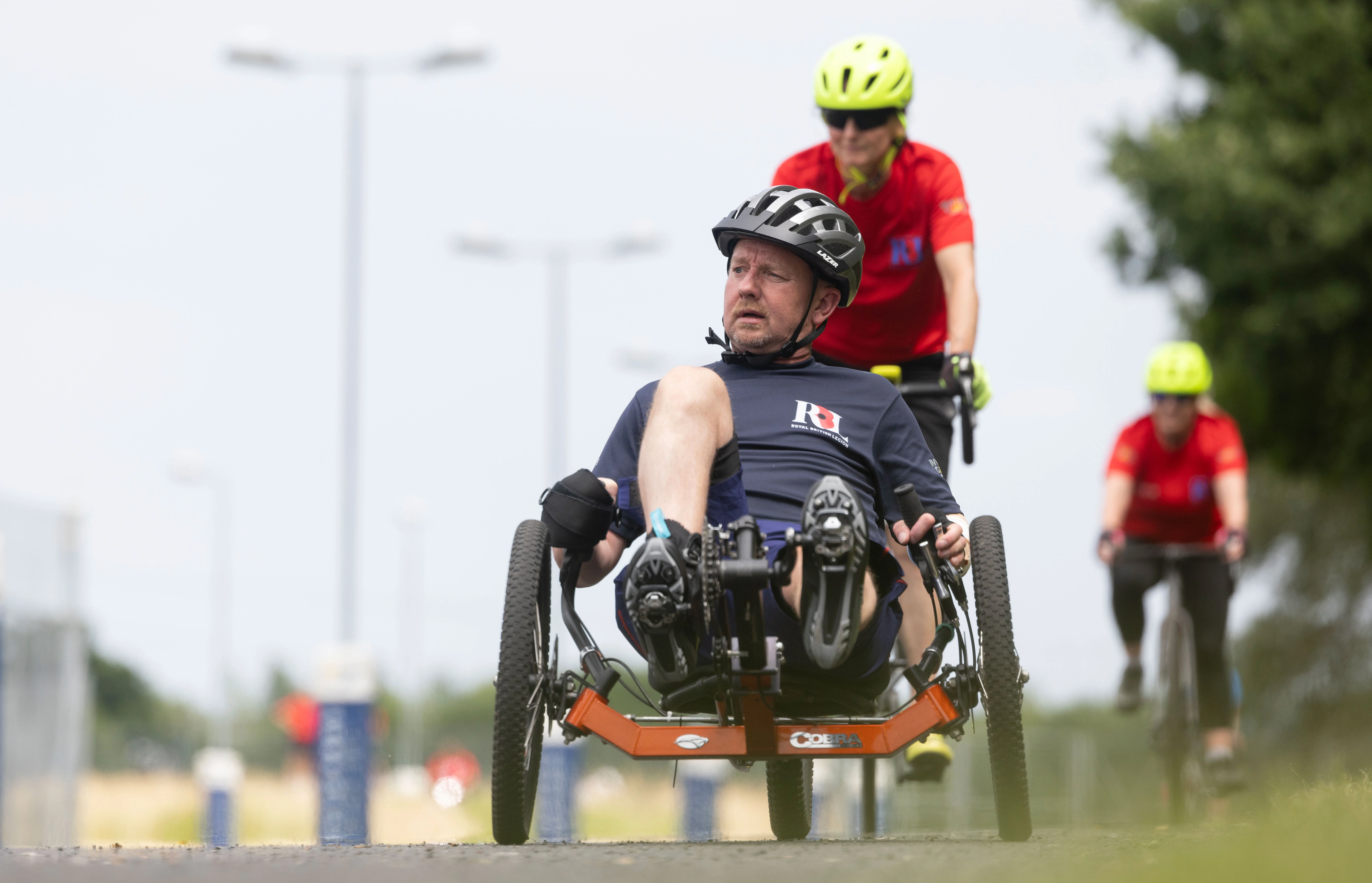 Al in a recumbent bike on a racing track. Behind him we can see another RBL competitor on a different adaptive bike. They are both wearing helmets and RBL t-shirts.
