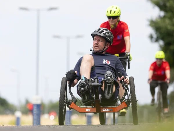 Al in a recumbent bike on a racing track. Behind him we can see another RBL competitor on a different adaptive bike. They are both wearing helmets and RBL t-shirts.