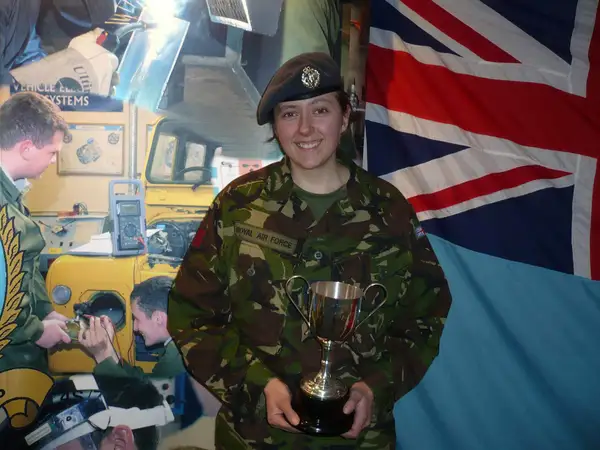 Liz in combat gear. She is smiling at the camera. She is holding a trophy in both hands. Behind her we can see a Union Jack strung up, as well as some Armed Forces personnel having a conversation in the background.