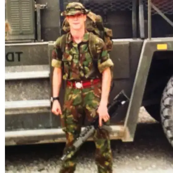 Steve in camouflage uniform holding a rifle in front of a military vehicle