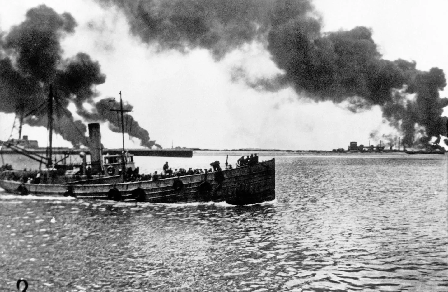 World war 2 Dunkirk evacuation. Black and white photo shows a small ship filled with soldiers on deck, moving left across calm water. Thick smoke rises from explosions on the horizon.