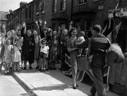 Neighbours wave and cheer as they watch a young soldier being welcomed home from the Second World War by his wife and baby