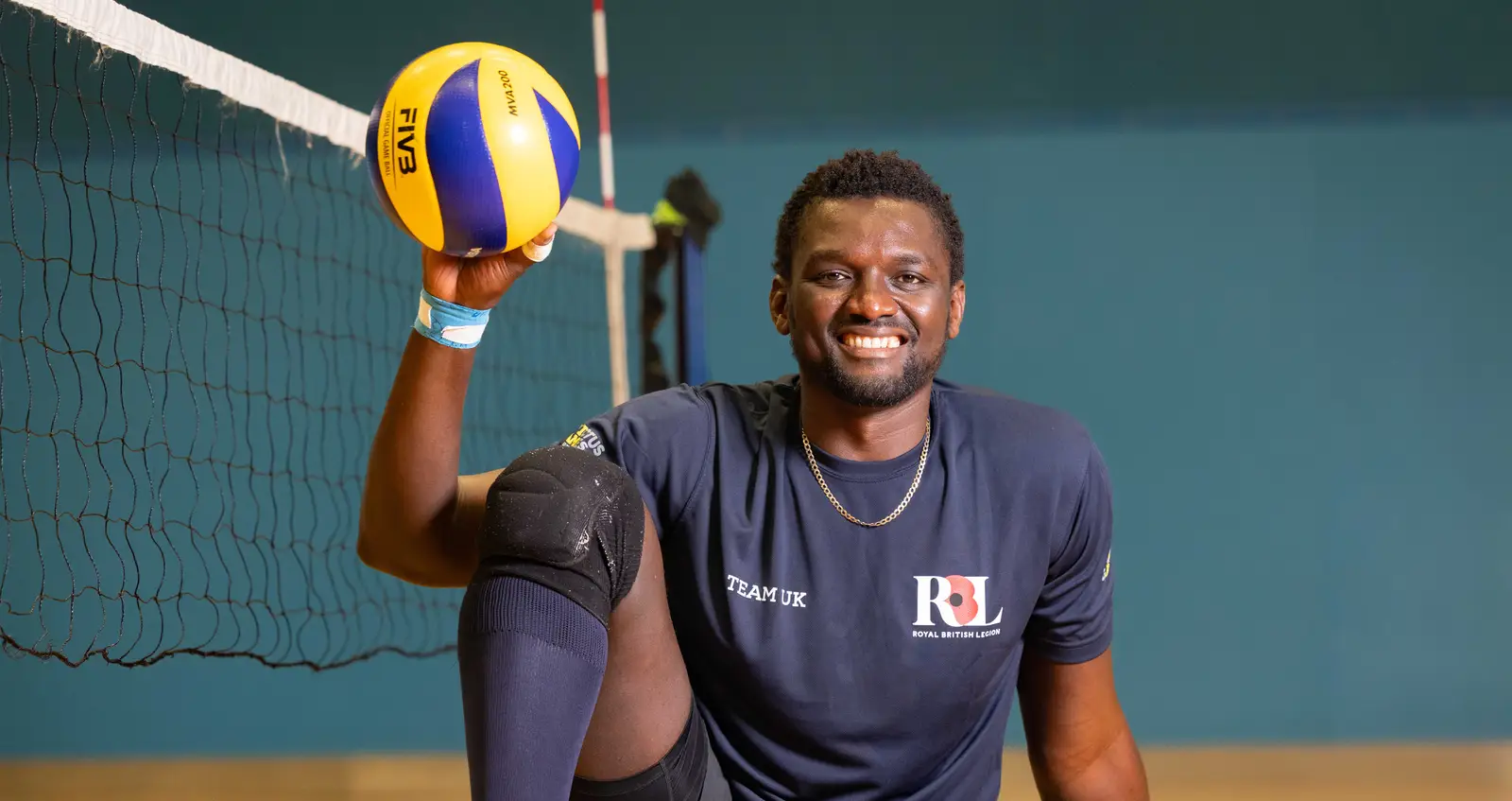 Emmanuel in his navy Invictus Games Team UK kit. He is sat on a volleyball court holding a volleyball up in the air in one of his hands. He is smiling.