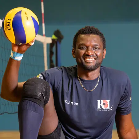 Emmanuel in his navy Invictus Games Team UK kit. He is sat on a volleyball court holding a volleyball up in the air in one of his hands. He is smiling.