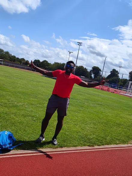 Emmanuel is wearing a red t-shirt and brown sports shorts. He is standing in the middle of a running track, his arms are outstretched and he looks happy. The sky above him looks bright and clear.