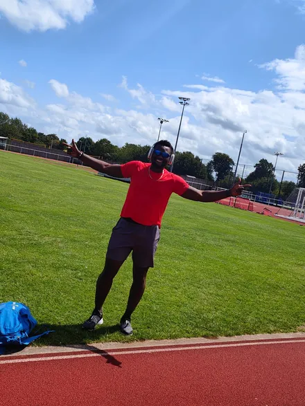 emmanuel image 5 Emmanuel is wearing a red t-shirt and brown sports shorts. He is standing in the middle of a running track, his arms are outstretched and he looks happy. The sky above him looks bright and clear.