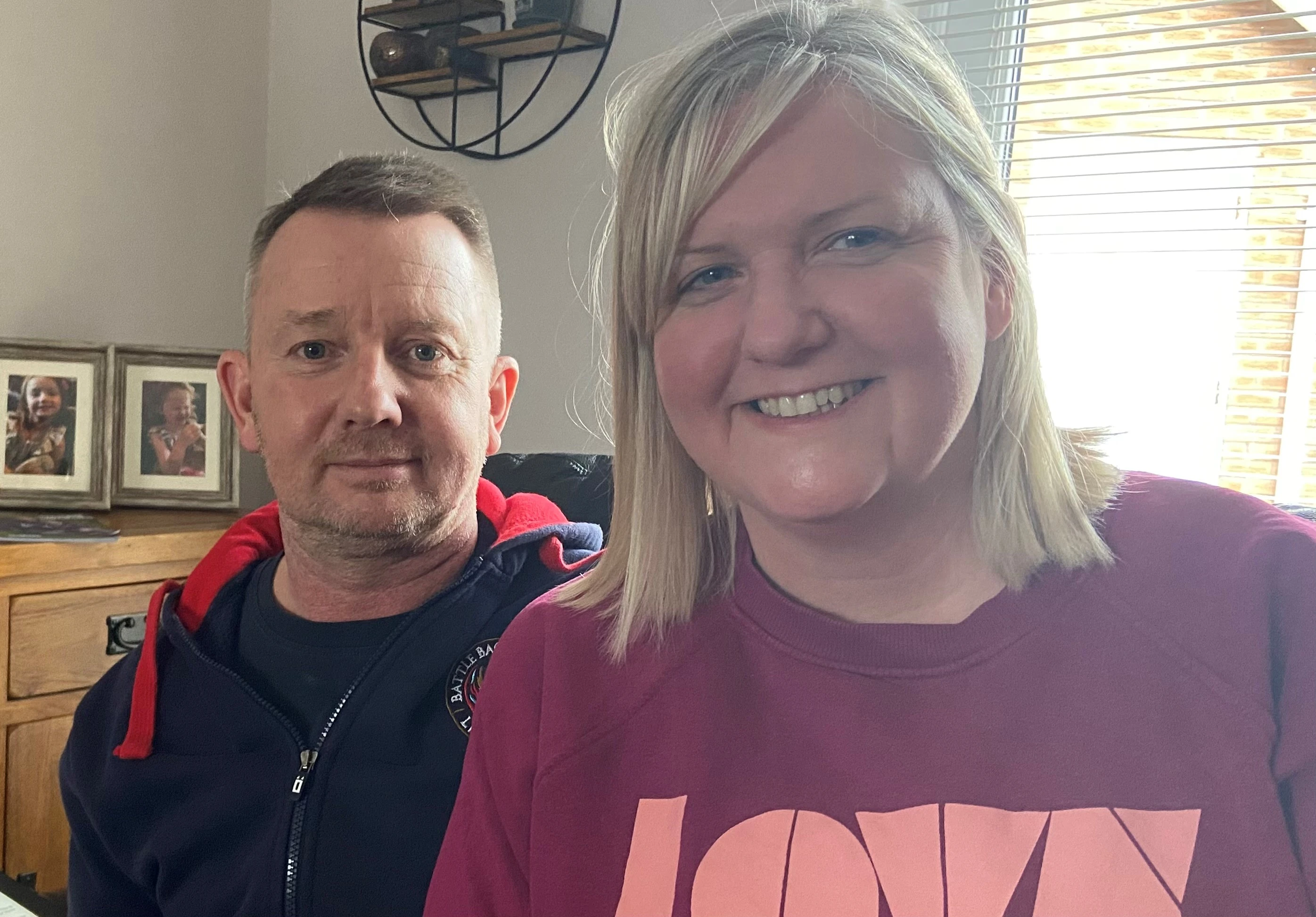 Al in dressed casually in a hoodie. He is sat next to his wife Gina, also dressed casually, who is taking the selfie. They are boht smiling. They appear to be at home, behind them we can see a chest of drawers with some framed photos on it.