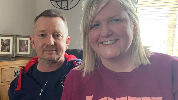 Al & Gina Dyer Al in dressed casually in a hoodie. He is sat next to his wife Gina, also dressed casually, who is taking the selfie. They are boht smiling. They appear to be at home, behind them we can see a chest of drawers with some framed photos on it.