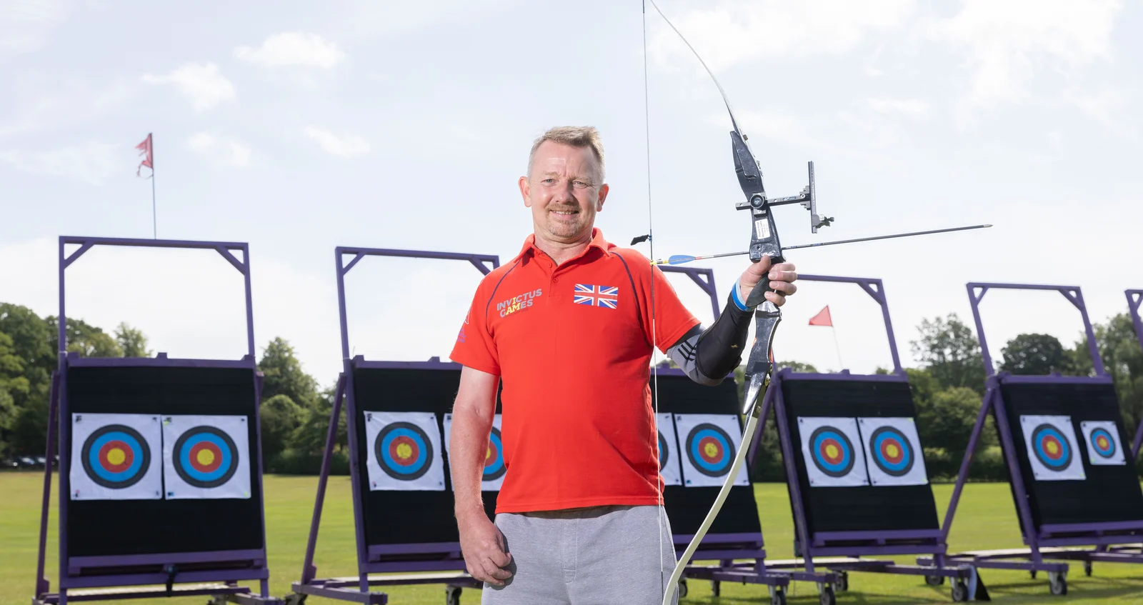 Al in a red Invictus Games t-shirt. He is looking straight at the camera smiling. In his left hand he's holding a crossbow . Behind him we can see some archery targets positioned on a green field.