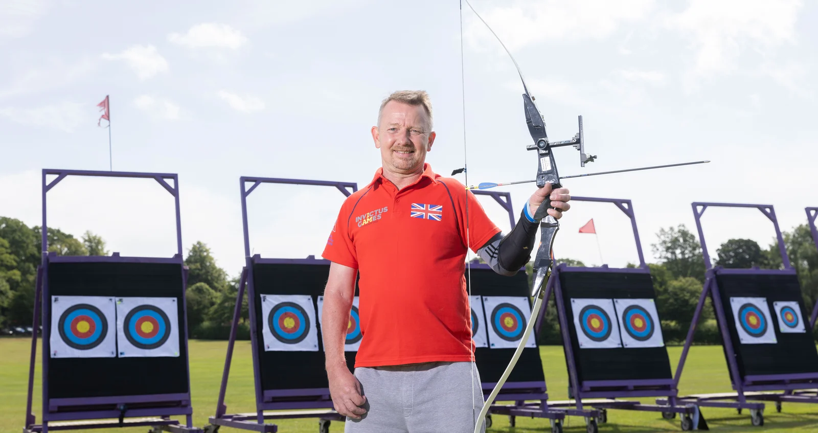 RBL_Invictus_Training_Camp-85 Al in a red Invictus Games t-shirt. He is looking straight at the camera smiling. In his left hand he's holding a crossbow . Behind him we can see some archery targets positioned on a green field.