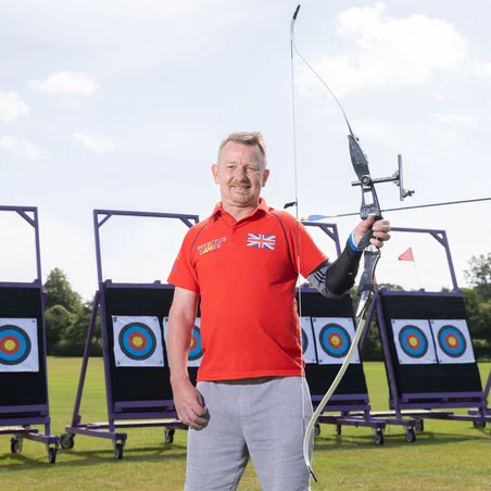 RBL_Invictus_Training_Camp-85 Al in a red Invictus Games t-shirt. He is looking straight at the camera smiling. In his left hand he's holding a crossbow . Behind him we can see some archery targets positioned on a green field.