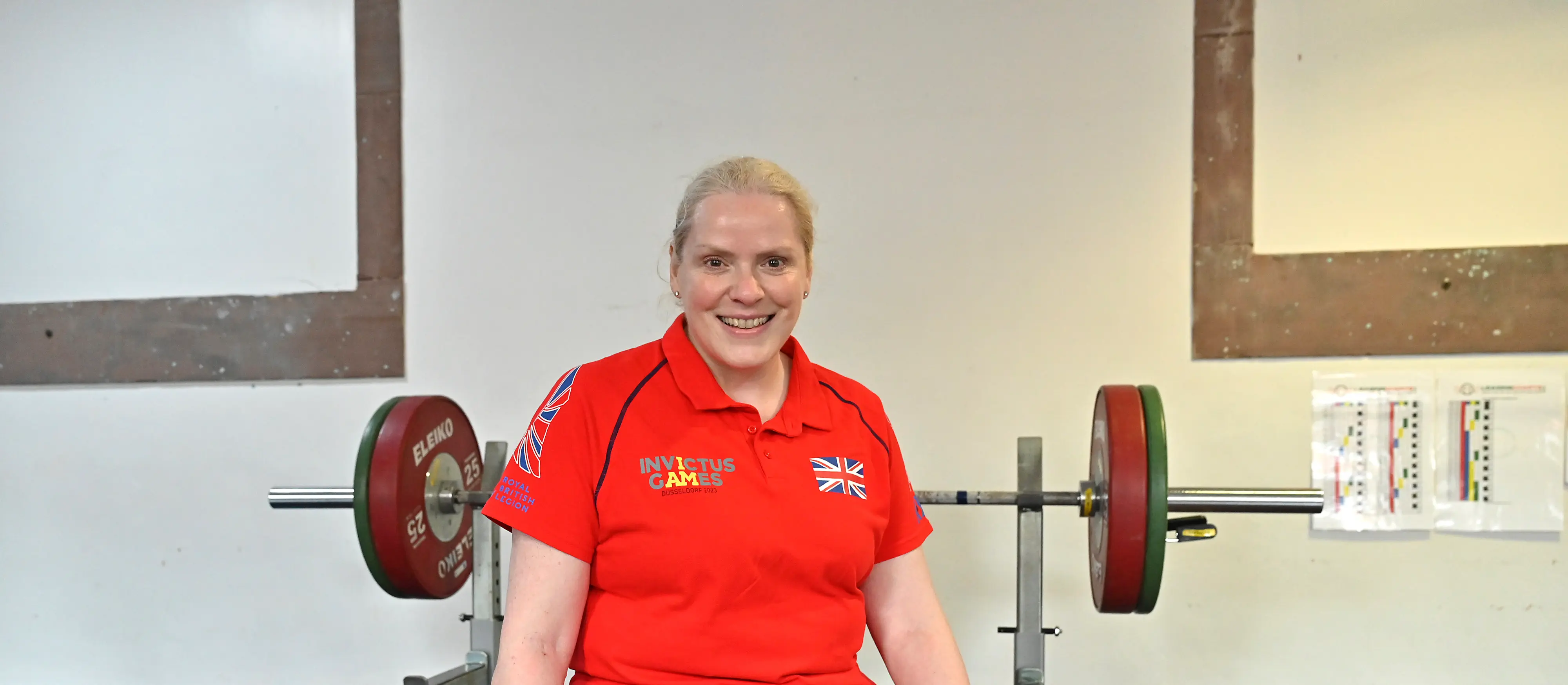 Amanda in a red Invictus Games Team UK polo shirt. There is a barbell with weights on it behind her.