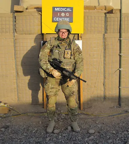 Photo of Amanda in full combat gear holding a gun, behind her we can see a yellow sign stuck on a makeshift wall which says Medical IG Centre.