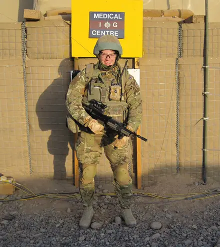 IMG-20230315-WA0000 Photo of Amanda in full combat gear holding a gun, behind her we can see a yellow sign stuck on a makeshift wall which says Medical IG Centre.
