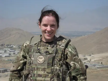 Close up photo of Amanda from the waist up. She is dressed in full combat gear and is smiling at the camera. Behind her we can see some scenic landscape views of what looks like a foreign country.