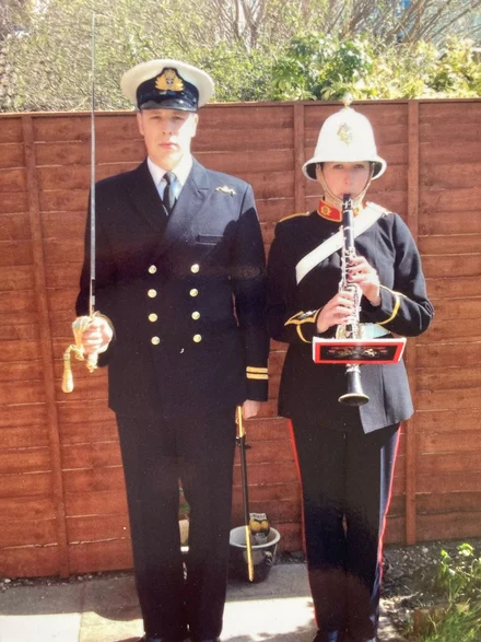Photograph of Becky and her brother James. They are both dressed in their military uniforms. James is standing to attention, holding a sword and Becky is standing to attention playing a clarinet. Behind them we can see a brown fence and some trees.