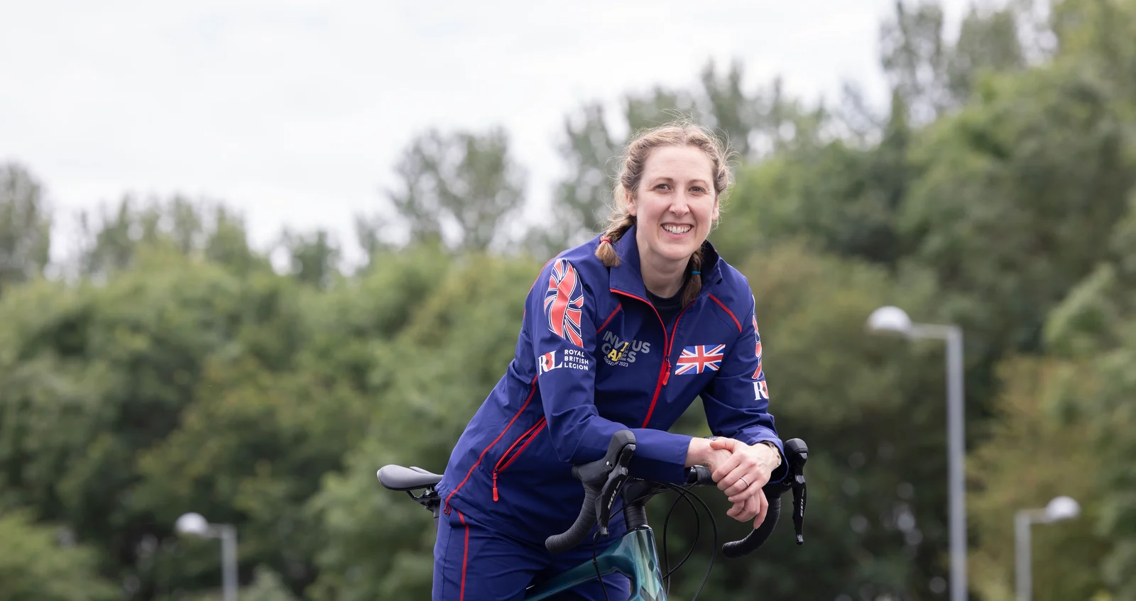 Becky leaning on her bike, smiling towards the camera. She is wearing a blue Invictus Games Team UK tracksuit and is smiling at the camera. Behind her we can see a background of trees.
