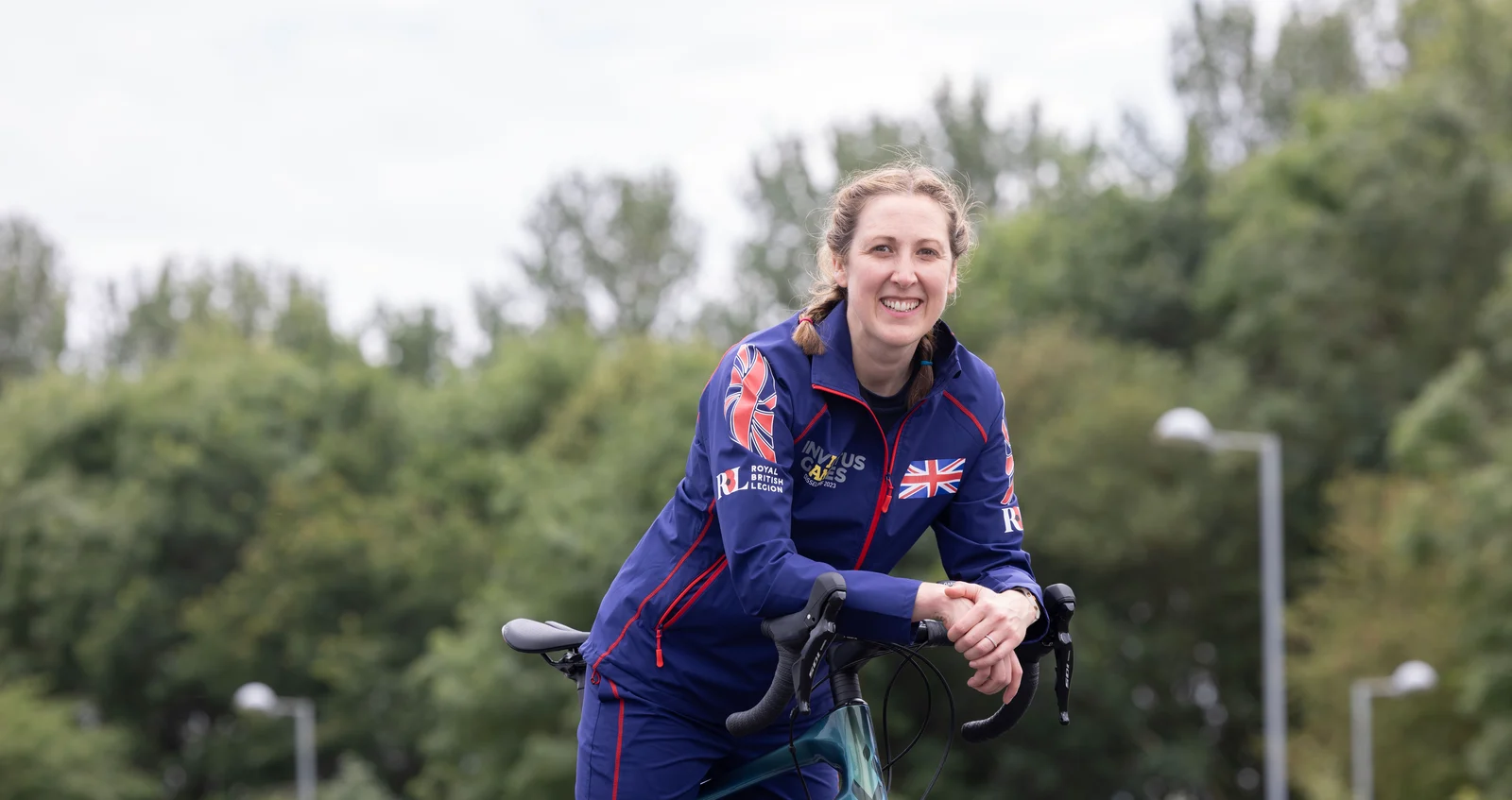 becky-image-3 Becky leaning on her bike, smiling towards the camera. She is wearing a blue Invictus Games Team UK tracksuit and is smiling at the camera. Behind her we can see a background of trees.