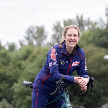 Becky leaning on her bike, smiling towards the camera. She is wearing a blue Invictus Games Team UK tracksuit and is smiling at the camera. Behind her we can see a background of trees.