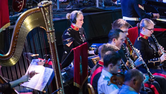 Becky playing the piano, dressed in her Royal Marines Band Service uniform. To the left of her we can see a big harp, and in front of her we can see other members of the band playing various instruments. The setting is a darkly lit concert hall.