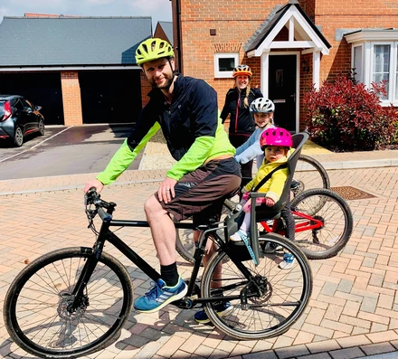 Becky standing in front of a red brick house, smiling and wearing a cycling helmet. In front of her we can see her partner and two children, also on bikes and wearing helmets.