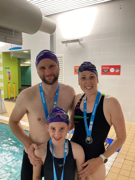 Becky, her partner and their child standing in front of them. They are all dressed in swimsuits with swim caps on and are also wearing blue medals around their necks. Behind them we can see a swimming pool and the entryway to some showers.