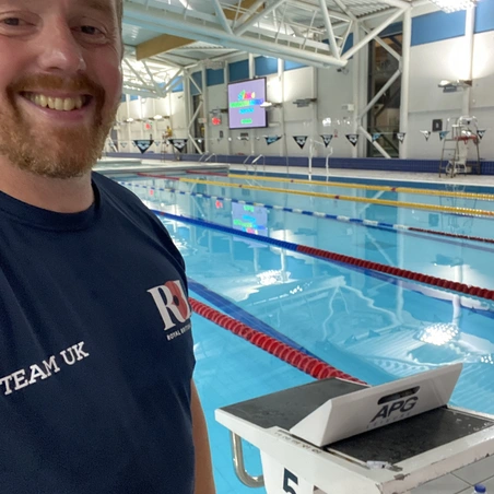 Charlie is wearing his Team UK Invictus Games navy t-shirt. He is taking a selfie and smiling. He is standing inside a leisure centre and behind him we can see a swimming pool.
