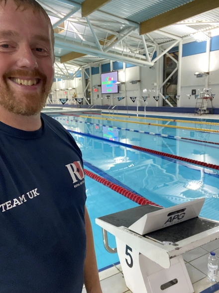 image00002 Charlie is wearing his Team UK Invictus Games navy t-shirt. He is taking a selfie and smiling. He is standing inside a leisure centre and behind him we can see a swimming pool.