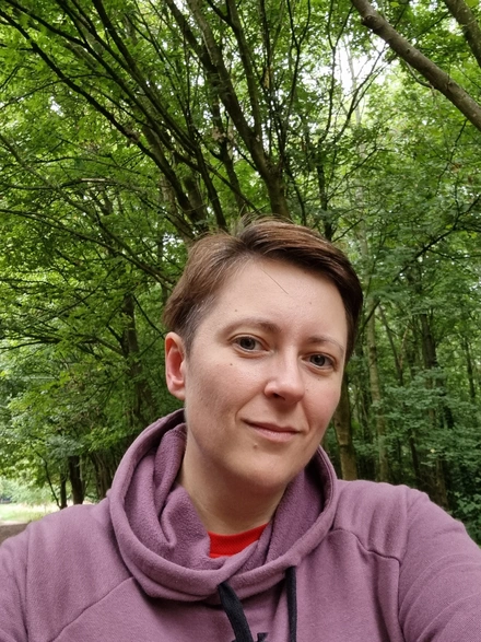 Selfie of Liz. She is wearing a purple hoodie and is smiling at the camera. Behind her we can see that she's standing in a forest.
