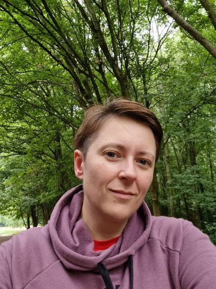 20220911_095903 Selfie of Liz. She is wearing a purple hoodie and is smiling at the camera. Behind her we can see that she's standing in a forest.
