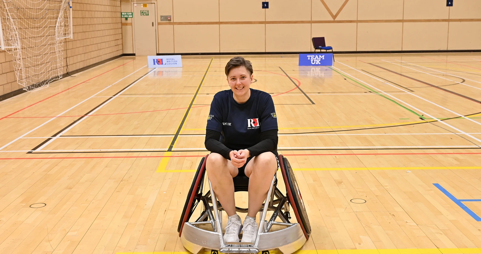 Liz in a wheelchair in a basketball court. She is facing the camera head on, wearing a navy Team UK RBL shirt. Behind her we can see some Team UK and RBL signs.
