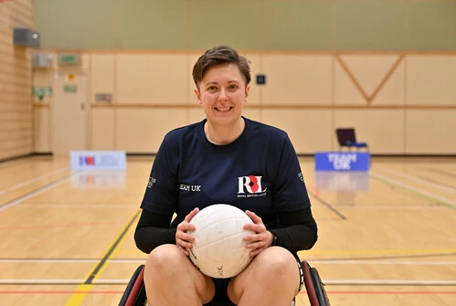 Liz in a wheelchair in a basketball court. She is facing the camera head on, wearing a navy Team UK RBL shirt. Behind her we can see some Team UK and RBL signs.