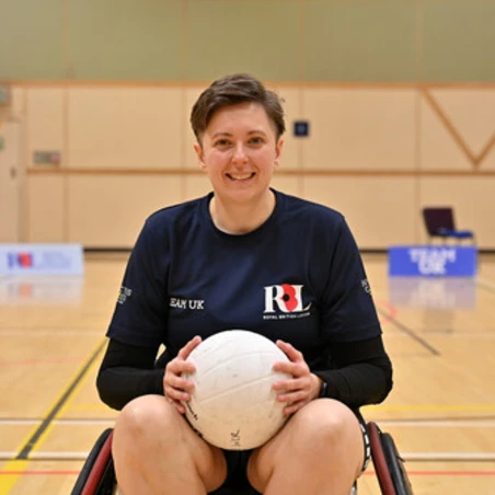 49919 Liz in a wheelchair in a basketball court. She is facing the camera head on, wearing a navy Team UK RBL shirt. Behind her we can see some Team UK and RBL signs.