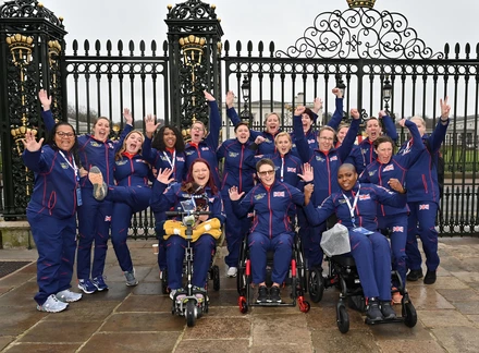 Group shot of Invictus Games female participants all wearing blue and red Team UK tracksuits. They all have their hands raised in a cheer as they wave at the camera. Behind them is a big black wrought iron gate. It’s a rainy and cloudy day outside.