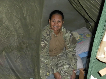 Tilly is smiling at the camera. She is wearing combat clothes. She appears to be sitting on a makeshift bed inside a green tent.