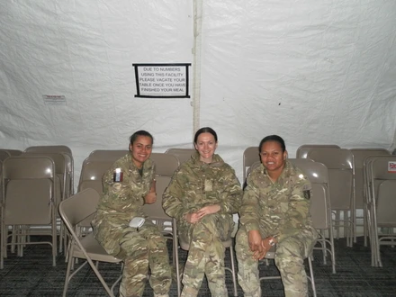 Tilly sat on chairs with two friends to her right. They are all sat on chairs inside what looks like a white tent, wearing combat gear.