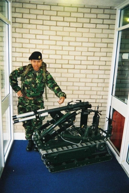 A young Tilly in combat gear standing next to a piece of weaponry. She's standing inside a corridor with glass doors either side of her and a brick wall behind her.