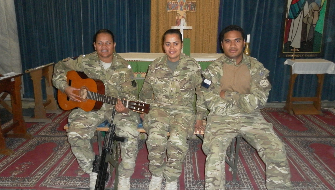 Tilly photographed with two friends inside a church. They are all wearing combat clothes and are sitting on a bench, smiling. Tilly is holding a guitar in her hands.