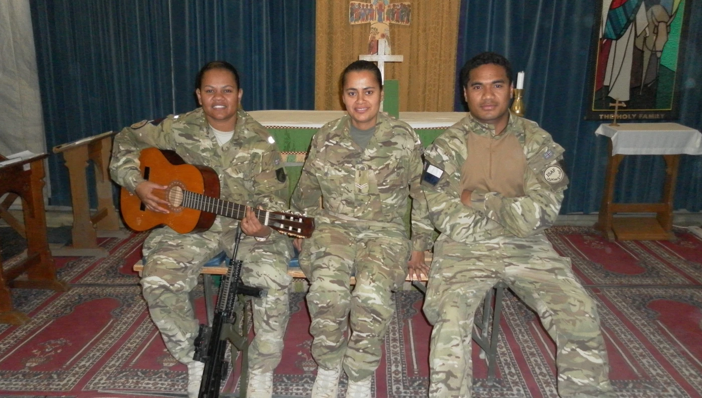 Tilly Tilly photographed with two friends inside a church. They are all wearing combat clothes and are sitting on a bench, smiling. Tilly is holding a guitar in her hands.